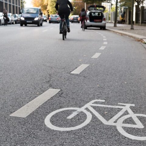 Ein Fahrradsymbol und eine gestrichelte Linie am Straßenrand machen auf die Radwegbenutzungspflicht aufmerksam. Weiter hinten auf der Straße fahren Autos und ein Radfahrer, der den Radweg nicht nutzen kann, weil weiter hinten ein Auto darauf parkt.