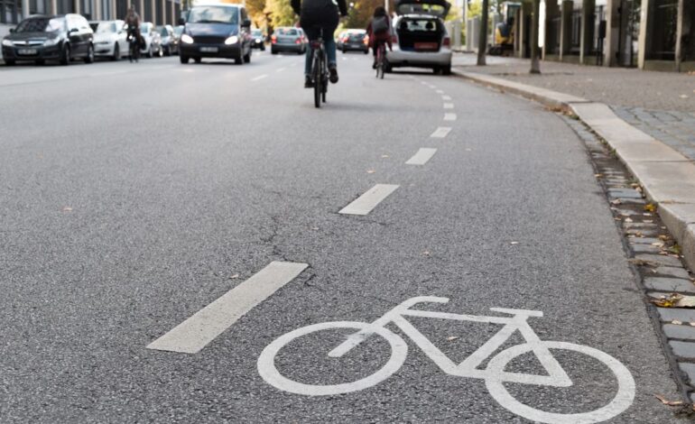 Ein Fahrradsymbol und eine gestrichelte Linie am Straßenrand machen auf die Radwegbenutzungspflicht aufmerksam. Weiter hinten auf der Straße fahren Autos und ein Radfahrer, der den Radweg nicht nutzen kann, weil weiter hinten ein Auto darauf parkt.