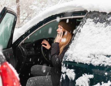 Frau in Winterkleidung sitzt bei offener Tür auf dem Fahrersitz eines Autos und telefoniert. Das Auto ist mit Schnee bedeckt und es schneit.