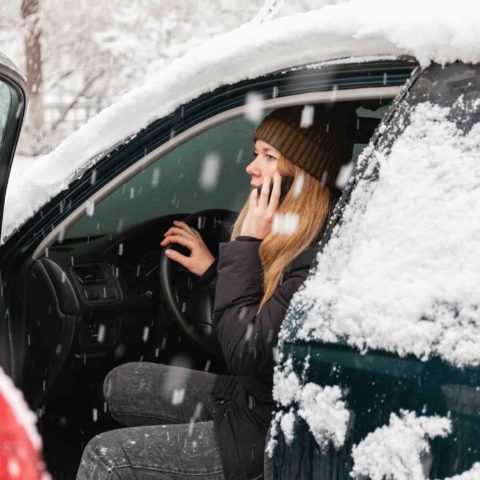 Frau in Winterkleidung sitzt bei offener Tür auf dem Fahrersitz eines Autos und telefoniert. Das Auto ist mit Schnee bedeckt und es schneit.