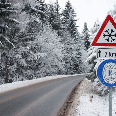 Straße durch einen verschneiten Wald. Verkehrsschild am Rand mit Schneeketten Symbol unter einem Warnschild für Schneefall für die nächsten 7 km