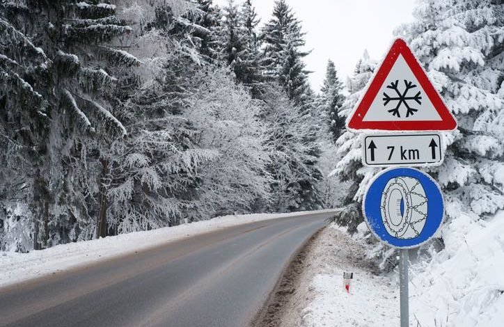 Straße durch einen verschneiten Wald. Verkehrsschild am Rand mit Schneeketten Symbol unter einem Warnschild für Schneefall für die nächsten 7 km