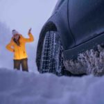 Auto im Schnee im Vordergrund, Fokus von unten auf Vorderreifen, Schnee hängt am Unterboden. Frau in Winterbekleidung im Hintergrund hebt ratlos eine Hand.