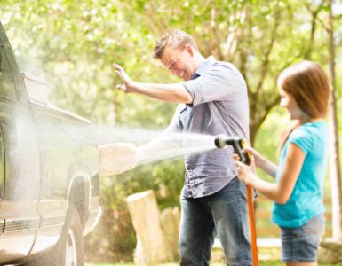 Vater und Tochter bei der Autowäsche. Tochter spritzt Wasser auf das Auto mit einem Gartenschlauch.
