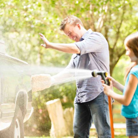 Vater und Tochter bei der Autowäsche. Tochter spritzt Wasser auf das Auto mit einem Gartenschlauch.