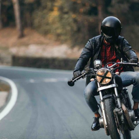 Motorradfahrer mit schwarzem Helm, Lederjacke und Jeans fährt auf einer Straße auf die Kamera zu.