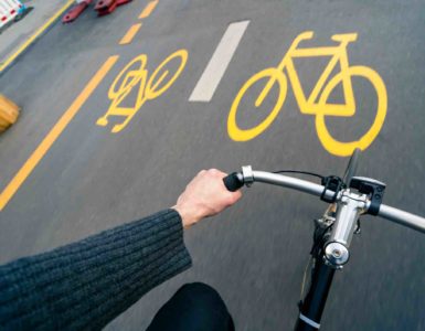Sicht von oben auf den Lenker eines Fahrrads mit der linken Hand des Fahrers. Im Hintergrund gelbe Markierung von Pop-up-Radwegen.
