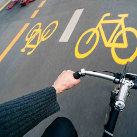 Sicht von oben auf den Lenker eines Fahrrads mit der linken Hand des Fahrers. Im Hintergrund gelbe Markierung von Pop-up-Radwegen.