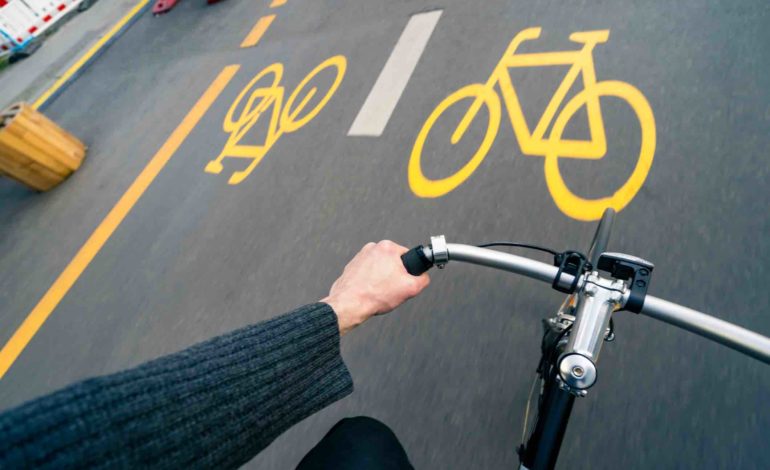 Sicht von oben auf den Lenker eines Fahrrads mit der linken Hand des Fahrers. Im Hintergrund gelbe Markierung von Pop-up-Radwegen.