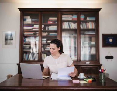 Frau mit Pferdeschwanz sitzt an einem Schreibtisch mit Laptop, Papierunterlagen, Kaffeetasse und Stiften. Im Hintergrund ist eine große Vitrine mit Büchern und Unterlagen.