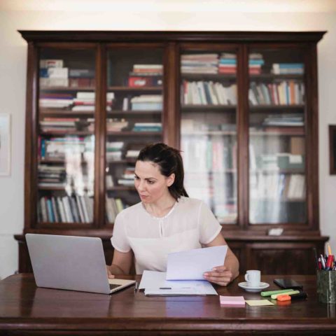 Frau mit Pferdeschwanz sitzt an einem Schreibtisch mit Laptop, Papierunterlagen, Kaffeetasse und Stiften. Im Hintergrund ist eine große Vitrine mit Büchern und Unterlagen.