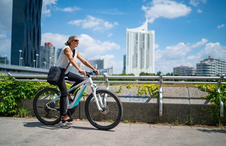 Frau mit Umhängetasche fährt auf einem E-Bike. Hochhäuser und blauer Himmel im Hintergrund.