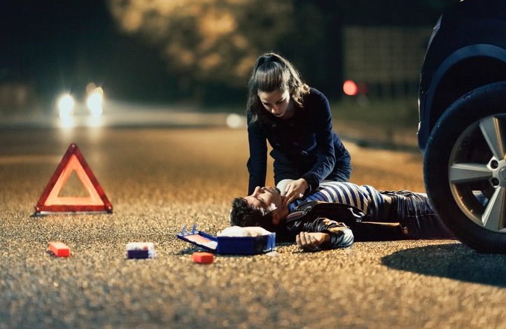 Frau wendet am Abend Erste Hilfe bei einem Mann an, der auf der Straße liegt. Daneben liegen der Verbandskasten und ein Warndreieck. Im Hintergrund sind Scheinwerfer zu sehen.
