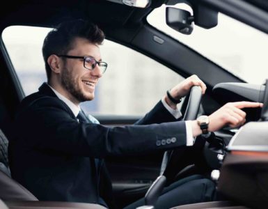 Mann mit Brille und Anzug im Auto mit einer Hand am Leckrad und einer Hand am Touchscreen im Armeturenbrett.
