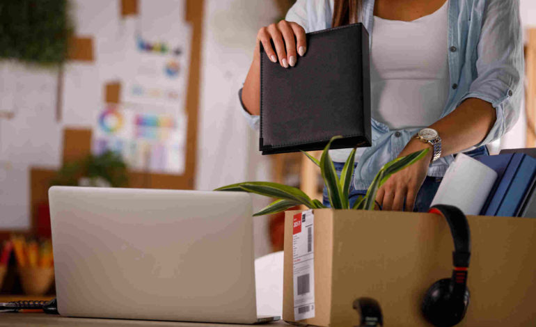 Frau packt Arbeitsunterlagen, Kopfhörer und eine Pflanze in einen Karton. Daneben steht ein aufgeklappter Laptop.