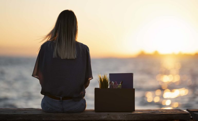 Junge Frau sitzt mit dem Rücken zur Kamera auf einer Mauer. Neben ihr steht eine Box mit Unterlagen, Schreibutensilien und einer Pflanze. Im Hintergrund ist ein Sonnenuntergang über dem Meer zu sehen.
