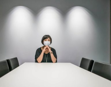 Vereins- und Gesellschaftsrecht: Eine Frau mit Mund-Nasen-Maske sitzt alleine an einem großen Tisch in einem Sitzungssaal. Die anderen vier Stühle sind leer.