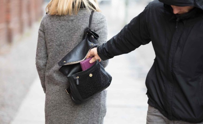 Ein Mann mit Kapuze über dem Gesicht stiehlt einer vorbeigehenden Dame das Portmonnaie aus ihrer Tasche.