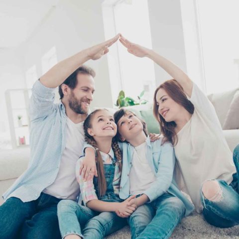 Eine lachende Familie sitzt auf dem Boden und zwei Personen halten jeweils eine Hand nach oben, zum Hausdach geformt.