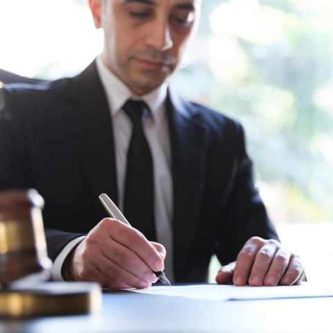 Legal-Tech-Gesetz: Anwalt sitzt am Schreibtisch und schreibt etwas auf ein Blatt. Im Vordergrund sieht einen Richterhammer.