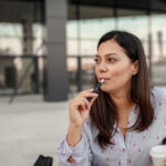 Eine Frau während der Raucherpause nach Arbeitsrecht, in einer Hand eine E-Zigarette, in der anderen einen Kaffeebecher.