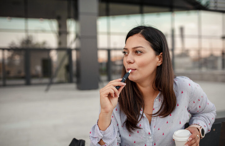 Eine Frau während der Raucherpause nach Arbeitsrecht, in einer Hand eine E-Zigarette, in der anderen einen Kaffeebecher.