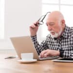 Älterer Mann sitzt vor einem Laptop am Tisch, eine Brille in der Hand. Davor stehen eine Kaffeetasse, eine Pflanze, ein Tablet und ein Smartphone.