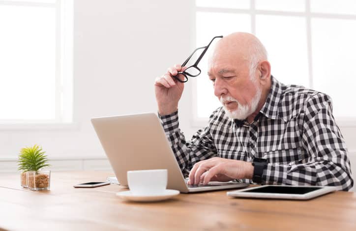 Älterer Mann sitzt vor einem Laptop am Tisch, eine Brille in der Hand. Davor stehen eine Kaffeetasse, eine Pflanze, ein Tablet und ein Smartphone.