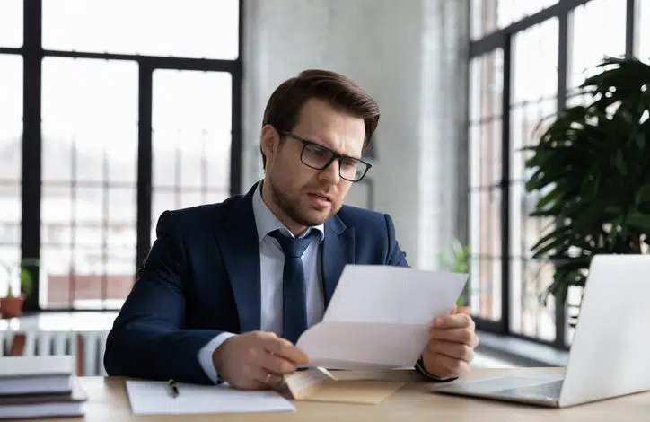 Mann mit Anzug, Krawatte und Brille liest mit besorgtem Gesichtsausdruck einen Brief am Schreibtisch. Auf dem Schreibtisch steht ein Laptop, im Hintergrund sind hohe Fenster.