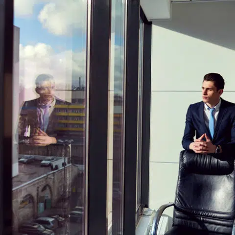 Geschäftsmann steht hinter einem leeren Lederstuhl und blickt nachdenklich aus dem Fenster.