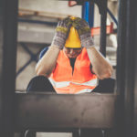 Ein Bauarbeiter mit orangefarbener Weste, gelbem Helm und Handschuhen sitzt auf einer Baustelle und hält sich frustriert die Hände an den Kopf.