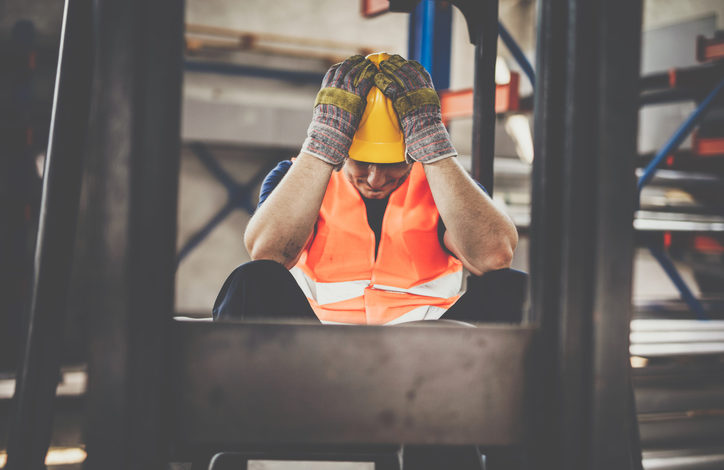 Ein Bauarbeiter mit orangefarbener Weste, gelbem Helm und Handschuhen sitzt auf einer Baustelle und hält sich frustriert die Hände an den Kopf.