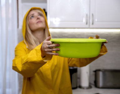 Eine Frau mit gelber Regenjacke und Kapuze schaut zur Decke. Von dort tropft Wasser in eine grüne Schüssel in ihren Händen. Im Hintergrund ist eine Küche zu sehen.