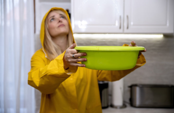 Eine Frau mit gelber Regenjacke und Kapuze schaut zur Decke. Von dort tropft Wasser in eine grüne Schüssel in ihren Händen. Im Hintergrund ist eine Küche zu sehen.