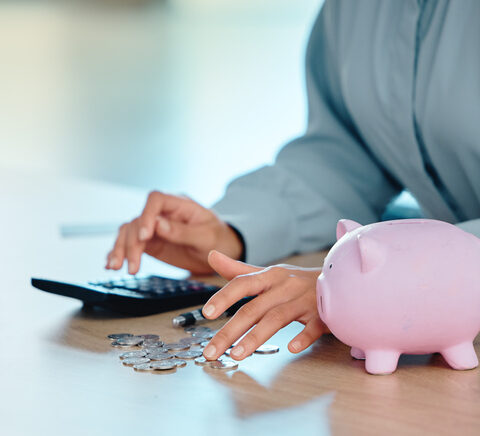 Frauenhände an einem Tisch berechnen die Insolvenz an einem Taschenrechner, daneben ein Sparschwein und mehrere Münzen.