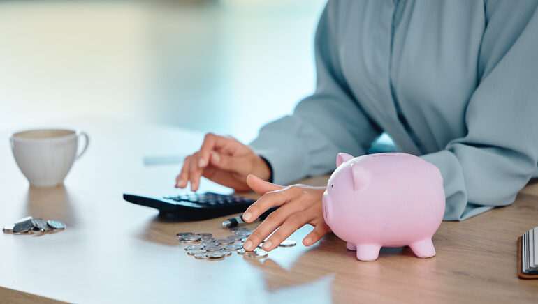 Frauenhände an einem Tisch berechnen die Insolvenz an einem Taschenrechner, daneben ein Sparschwein und mehrere Münzen.