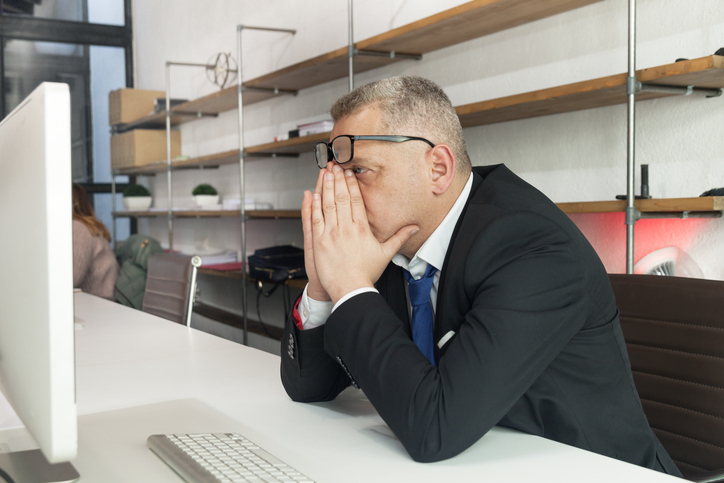 Ein Mann in Anzug und Krawatte, der während der Insolvenzbekanntmachung vor dem Computer sitzt. Er hält die Hände vors Gesicht und schiebt dabei seine Brille hoch. Im Hintergrund mehrere leere Regale.