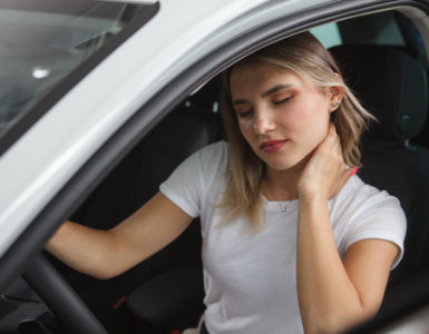 Junge blonde Frau mit weißem Shirt sitzt auf dem Fahrersitz eines Autos und reibt sich den Nacken.