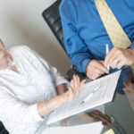 Eine Frau sitzt mit einem Mann am Tisch vor einem Rechtsdokument und hebt die Hand zum Widerspruch.