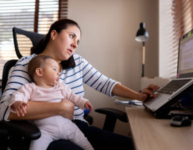 Mutter sitzt am Schreibtisch, auf dem Schoß ihr Baby. Zwischen Ohr und Schulter klemmt ihr Smartphone, mit einer Hand bedient sie ein Notebook.