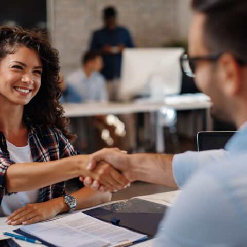 Junge Frau sitzt vor einem Schreibtisch und zeigt ihre Zustimmung durch konkludentes Handeln, indem sie lächelnd die Hand eines Mannes mit Brille auf der anderen Seite schüttelt.