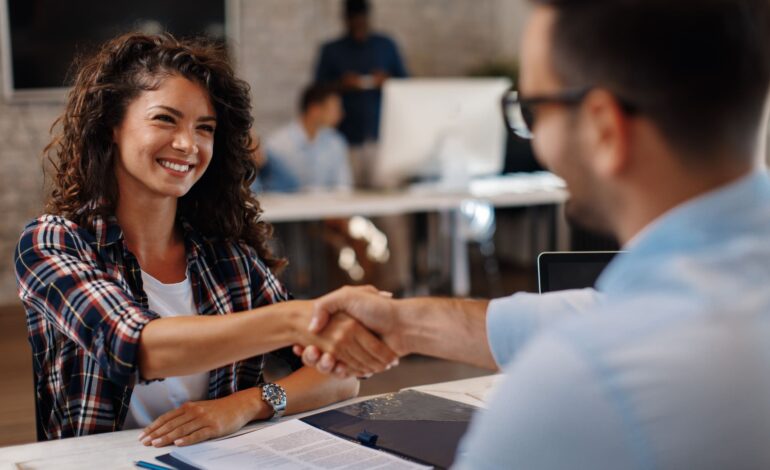 Junge Frau sitzt vor einem Schreibtisch und zeigt ihre Zustimmung durch konkludentes Handeln, indem sie lächelnd die Hand eines Mannes mit Brille auf der anderen Seite schüttelt.