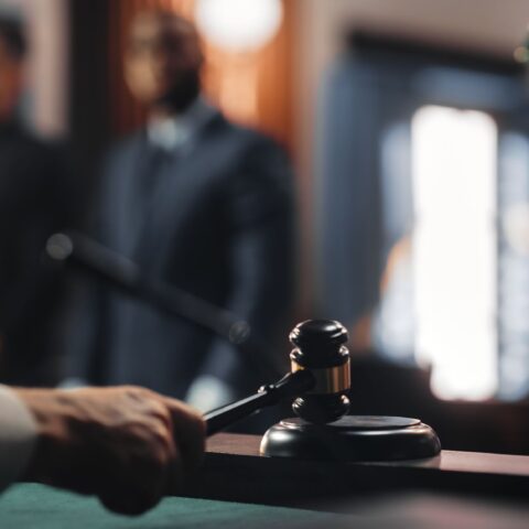 Die Hand eines Richters, der einen Richterhammer auf das Pult schlägt. Dahinter verschwommen zwei Personen, die das Schweigerecht anwenden.