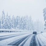 Schneebedeckte Fahrbahn und links und rechts zugeschneite Bäume. Ein Auto fährt auf der Straße.