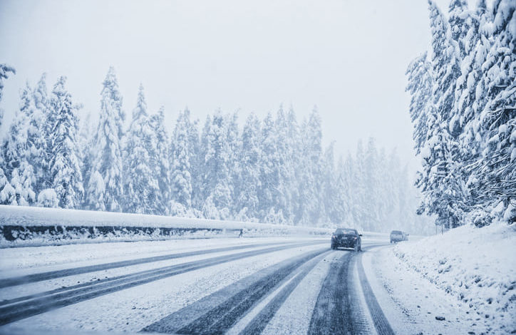 Schneebedeckte Fahrbahn und links und rechts zugeschneite Bäume. Ein Auto fährt auf der Straße.