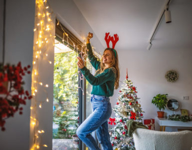 Eine junge Frau steht vor dem Fenster und hängt eine beleuchtete Lichterkette auf. Im Hintergrund ist weihnachtlich geschmückt.