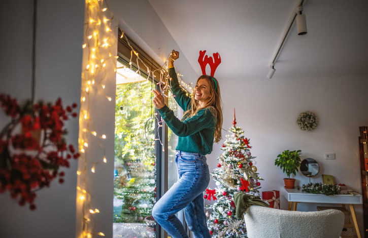 Eine junge Frau steht vor dem Fenster und hängt eine beleuchtete Lichterkette auf. Im Hintergrund ist weihnachtlich geschmückt.