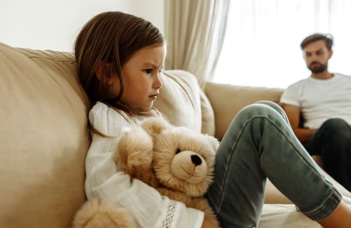 Auf einem Sofa sitzt ein trauriges Mädchen mit Teddybär im Arm. Dahinter entfernt ihr Vater. Es herrscht eine große Distanz.