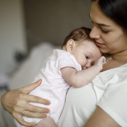 Eine junge Frau sitzt während der Elternzeit bei Adoption auf dem Sofa und hat ein Baby auf ihrer Brust liegen.
