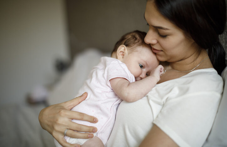 Eine junge Frau sitzt während der Elternzeit bei Adoption auf dem Sofa und hat ein Baby auf ihrer Brust liegen.